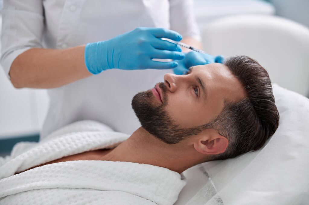 Lady cosmetologist holds syringe with filler above forehead of middle aged patient in salon