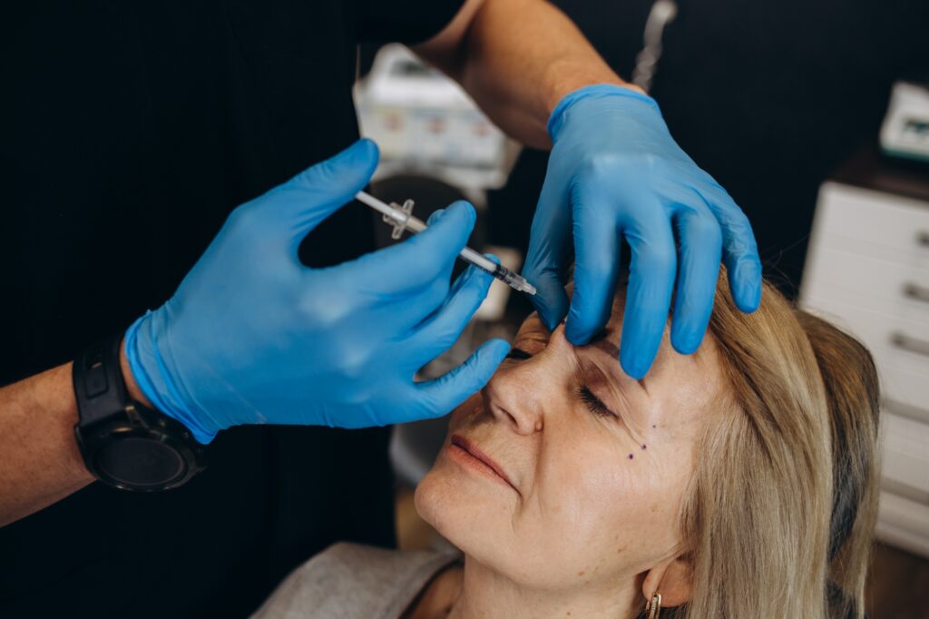 Plastic surgeon in protective gloves injecting face filler to senior woman over light studio
